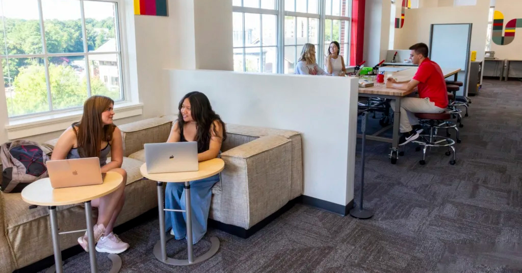 Two students seated in foreground and three in background with laptops in the new Makerspace in front of windows