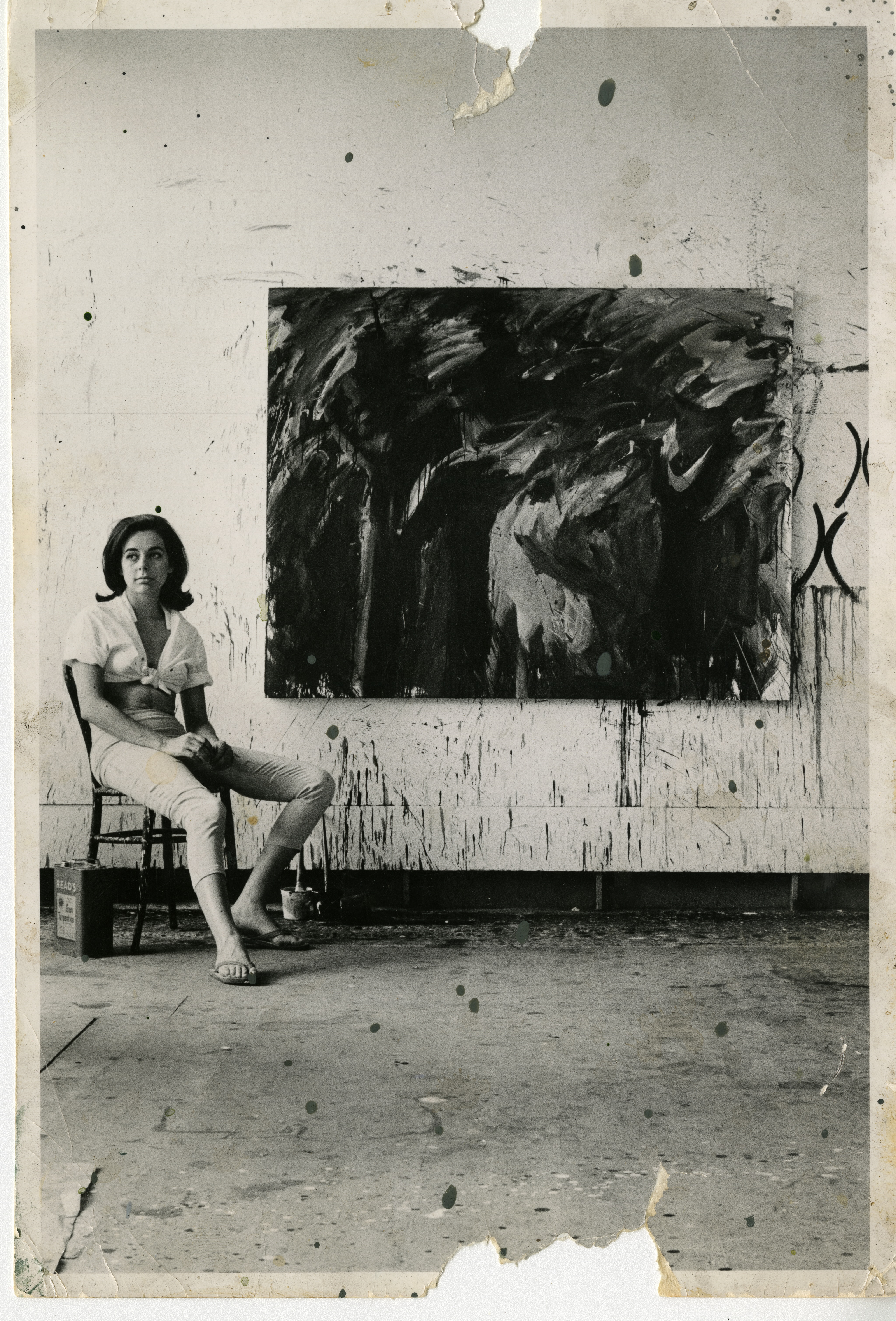 Nancy Ellison in a ripped black and white photo sitting in a chair beside an art piece