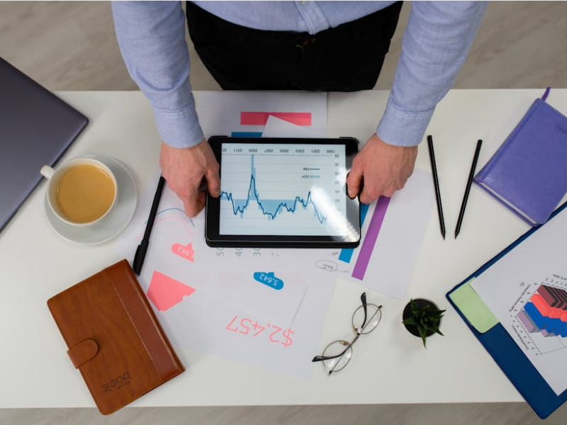 Person's hands on table gripping tablet with data visualization or graph on a messy desk with coffee cup, notebook, notes, glasses and pens