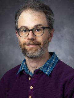 Bearded man with glasses wearing a blue shirt under a purple buttoned-up cardigan.