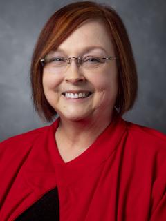 Woman with chin length bobbed red hair. She's wearing glasses and a red jacket over a black shirt.