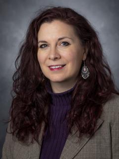 Woman with long dark red hair wearing a purple sweater under a tan blazer.