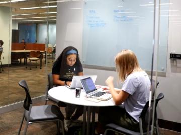 Two female undergrads sit at a round table, working on their laptops