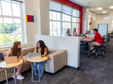 students seated in front of windows in foreground with laptops while several more students appear in the background at a higher table in the makerspace